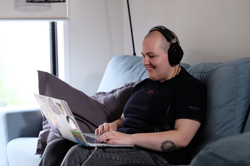 Jay sits on a couch with a laptop on their lap and earphones on.