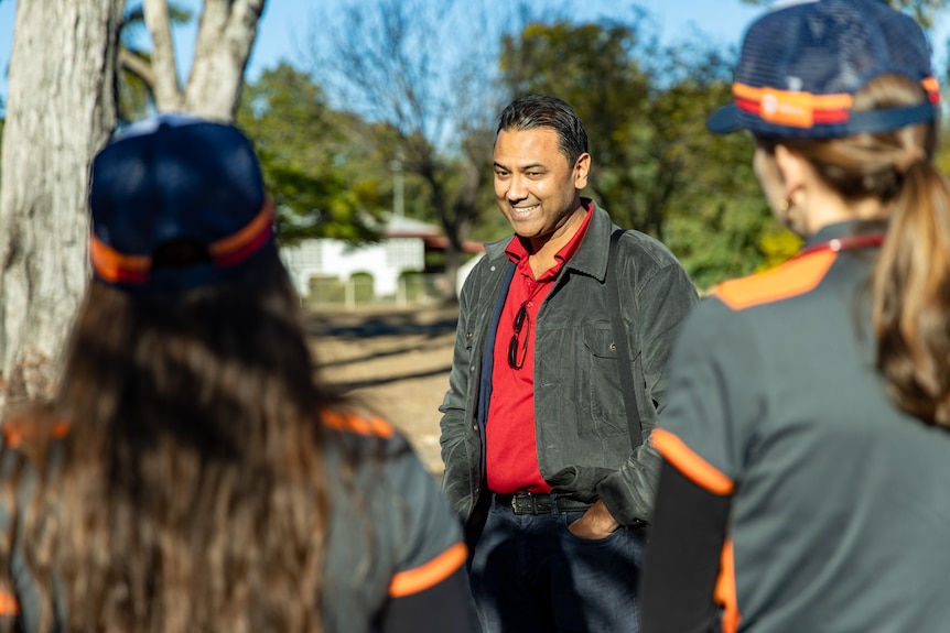 A man in a red shirt and black jacket stands with his hands in his pockets in front of a group of people.