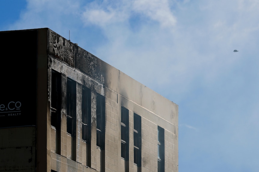 A grey building with large windows, the left side is charred and burnt. On the right a small drone is pictured in the air.