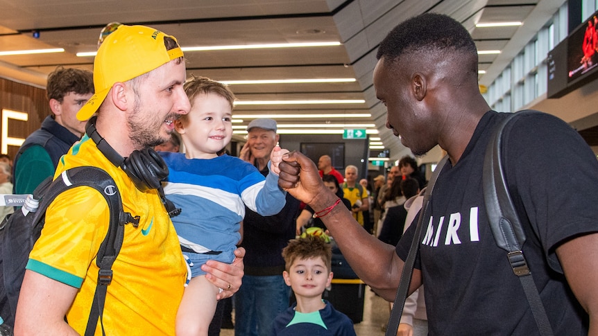 Thomas Deng fistbumps a toddler being held by his father, who is wearing Socceroos gear. 