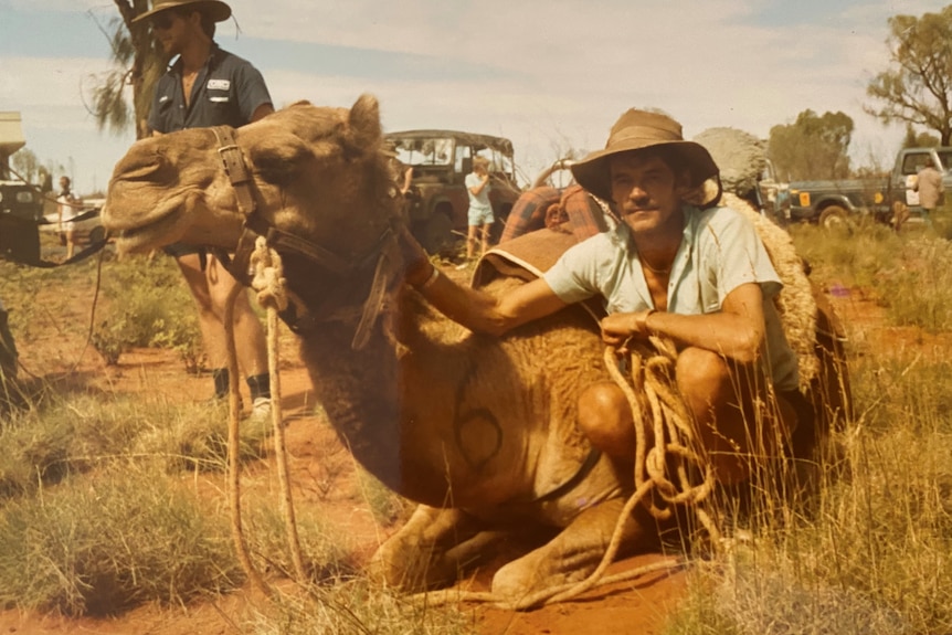 Man wearing wide-brimmed hat and short and t-shirt crouches down with sitting camel, in red dirt.
