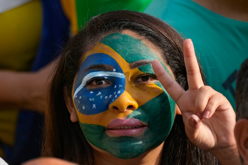A dark-haried woman with Brazliian flag face paint gives a peace sign