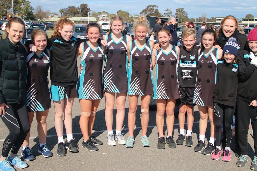 Team of young girls with coaches and Zac standing in a line, they wear and black uniform with blue sides