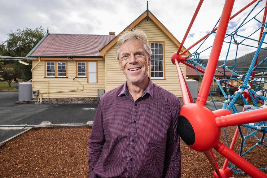 Man stands in front of school playground with weatherboard building in background.