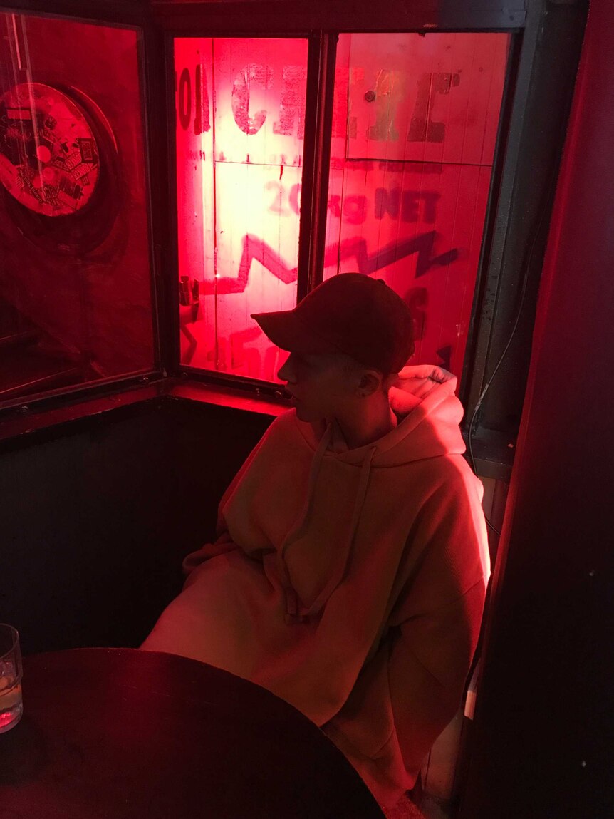 A man wears a hoodie and a cap sitting in a bar surrounded by red light.