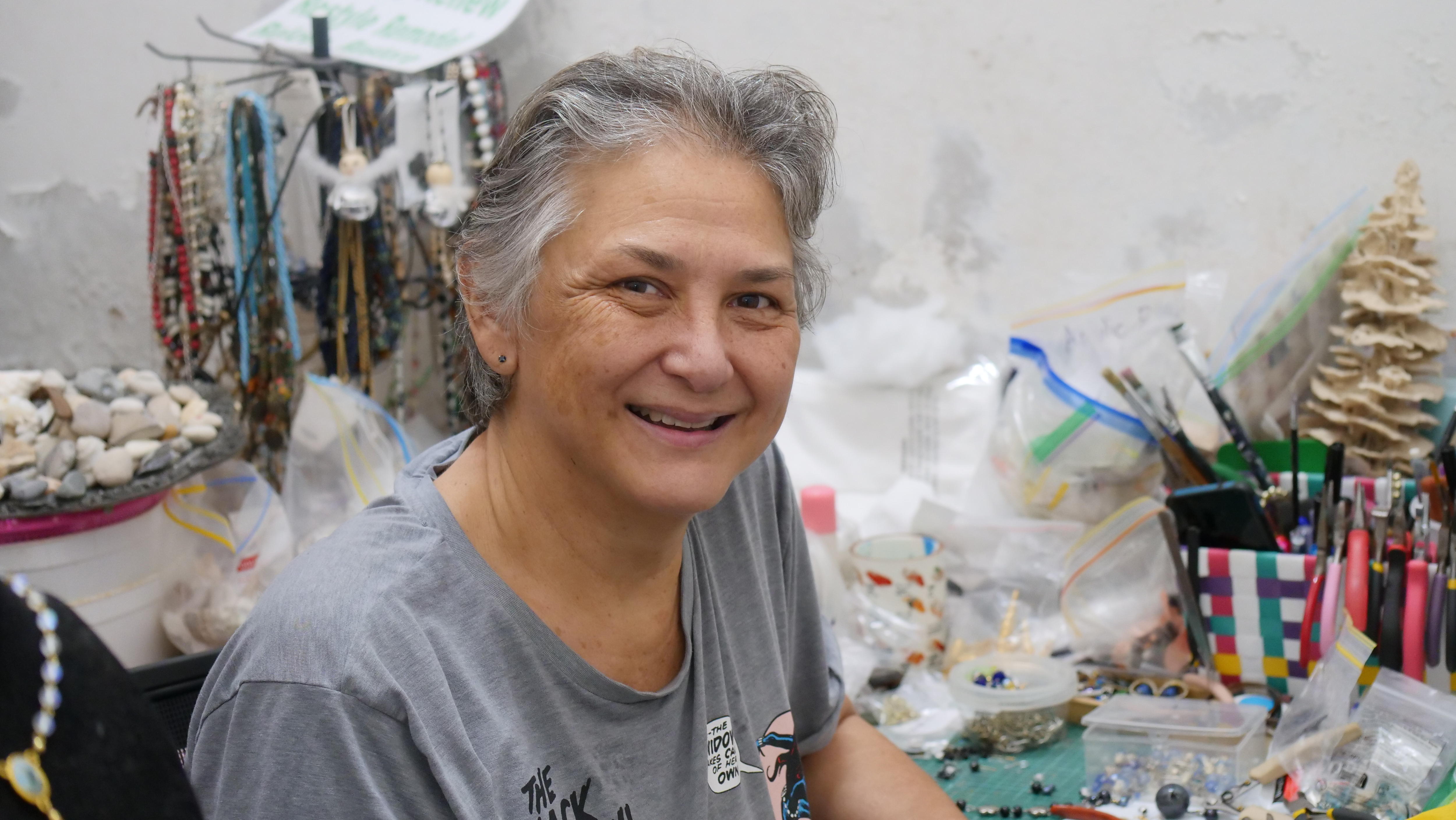 Uma mulher sorri diante de uma mesa cheia de materiais de arte. 