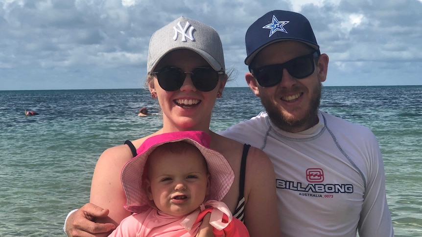 A young woman and man wearing caps and smiling on the beach. The woman is holding a baby wearing a bright pink hat