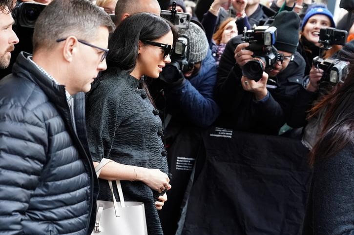 Meghan Markle wears a black coat and sunglasses and holds a grey bag as she is surrounded by paparazzi.