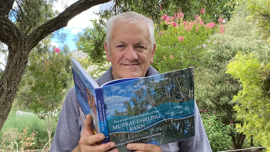 Adrian Wells holds a copy of his book while sitting in a garden.