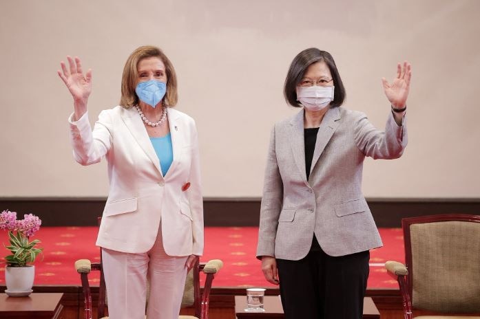 Two women in pant suits, wearing masks.  The one on the left is waving with her right hand from her.  The other woman with her left hand