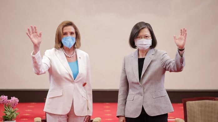 Two women in pant suits, wearing masks. The one on the left is waving with her right hand. The other woman with her left hand