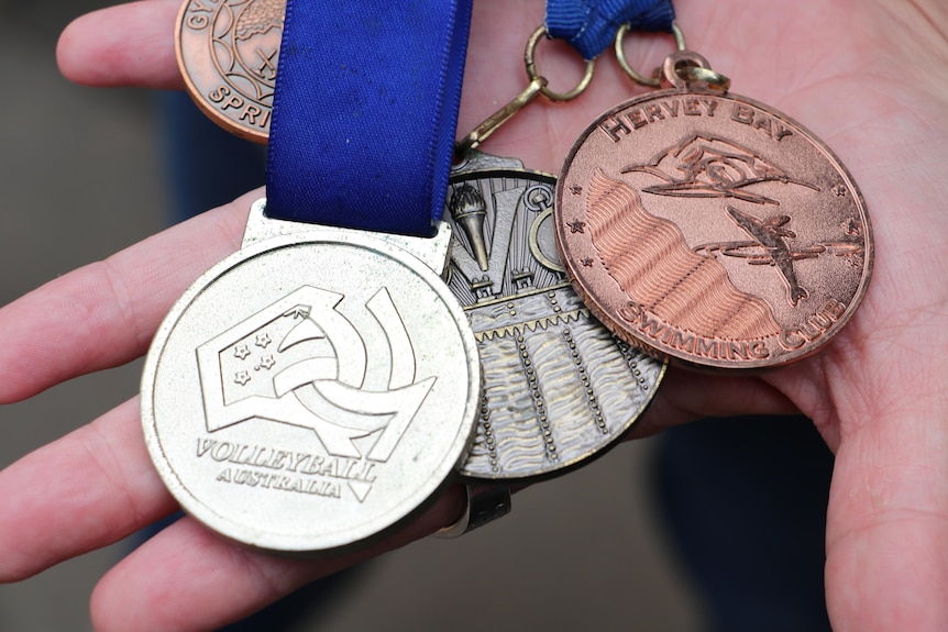A hand holds three sports medals, one is silver, one has a blue band one is brass
