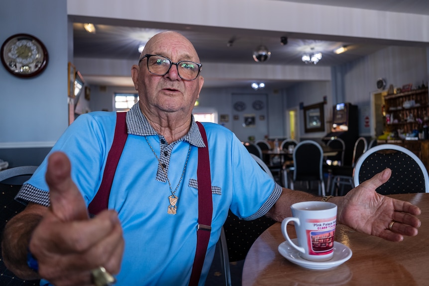 A man in a blue polo shirt and red suspenders sits at a table with a cup of tea