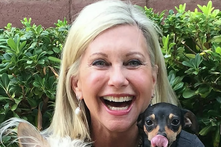 Dame Olivia Newton-John smiles as she hugs two small dogs in a garden.