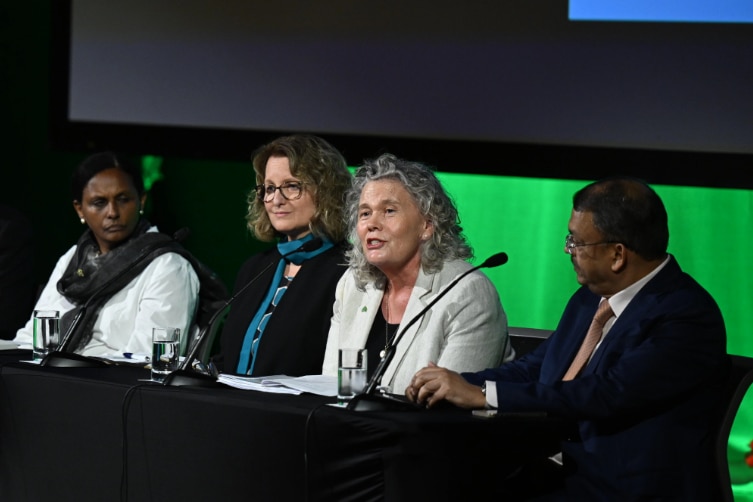 wide shot of six people seated behind a black table with microphones on a stage with a screen in the background