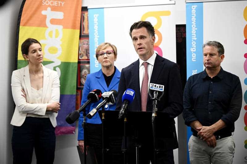 The premier speaks at a press conference with two women standing to his right and one man standing to his left.