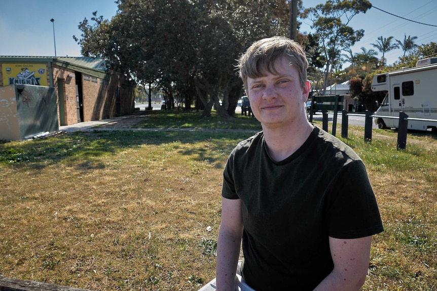 Dion Galea sits on a fence in the sunshine, smiling at the camera.