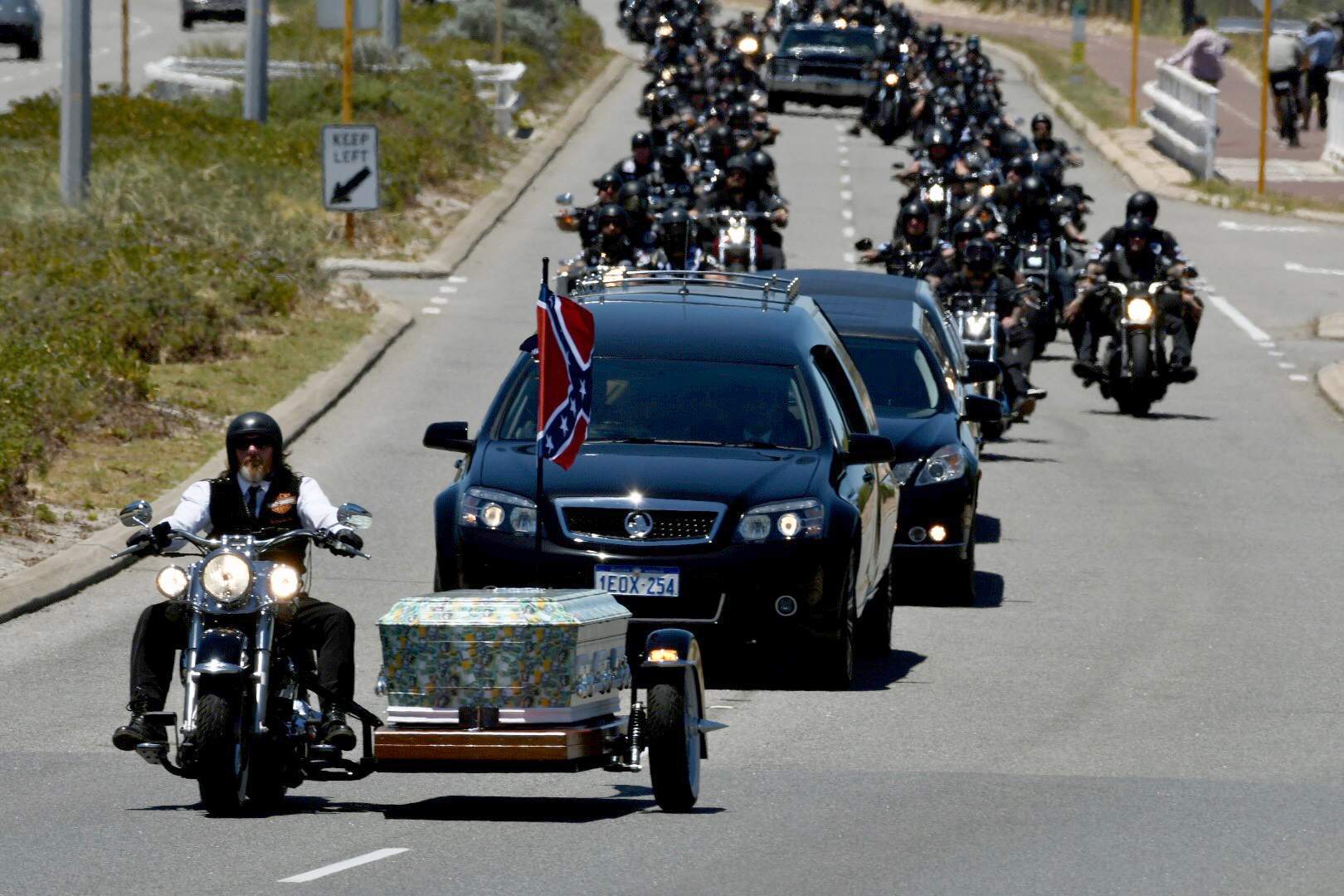 A man on a Harley rides in front of the coffin, with dozens of bikies riding behind it.