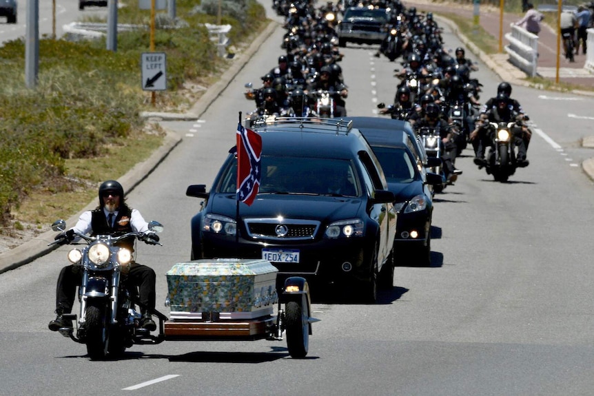 A man on a Harley rides in front of the coffin, with dozens of bikies riding behind it.