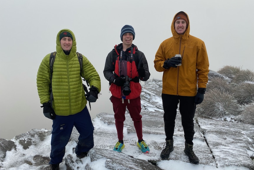 Three smiling young men standing on the peak of a snowy mountain.