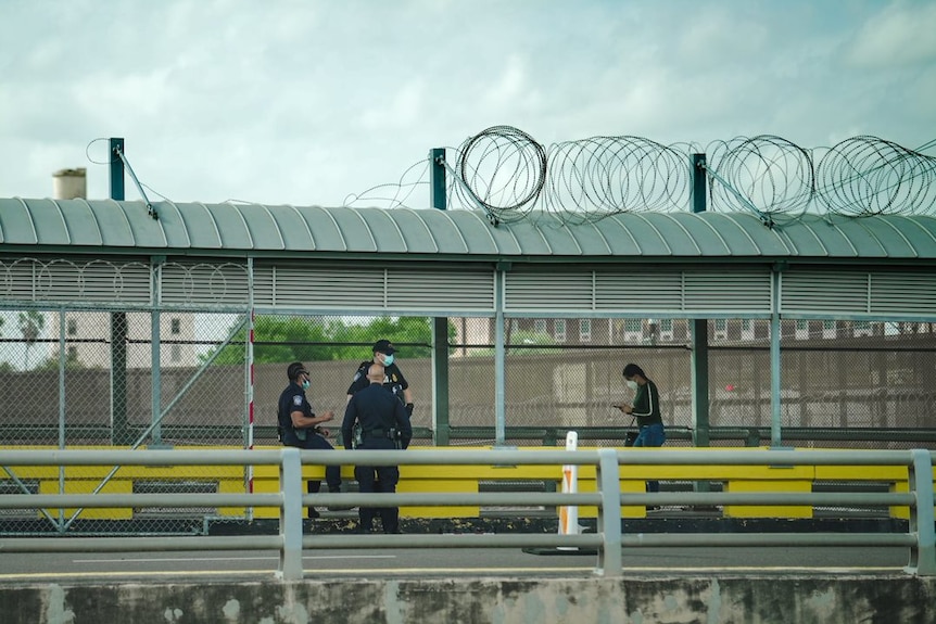 Three men dressed in dark blue uniform stand outside a fence as a person walks by looing at a phone.