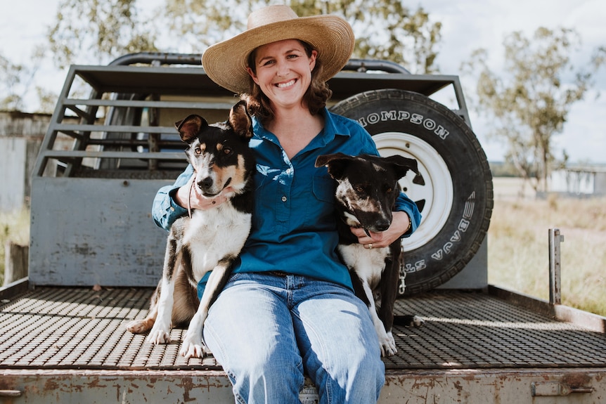 A woman sits on the back of a ute tray with two dogs