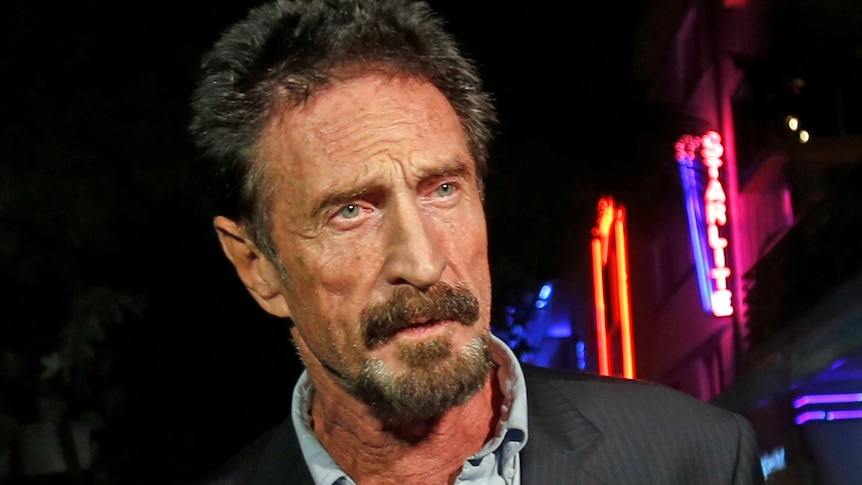 John McAfee in a suit looking right of screen with neon signs in the background