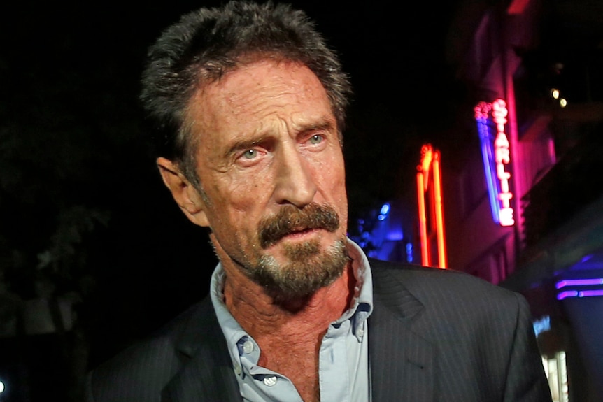 John McAfee mira a la derecha de la pantalla con etiquetas de neón en el fondo disfrazado