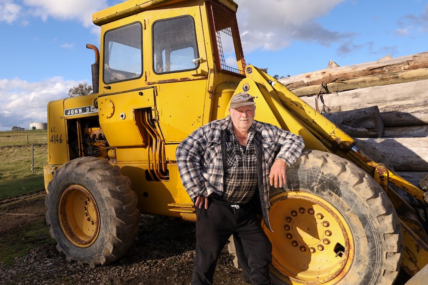 Older man in cap and flannelette shirt poses in front of big yellow dozer.