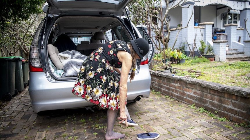 A woman in a dress and hat stands by a car in the driveway of a house while she puts her shoes on.