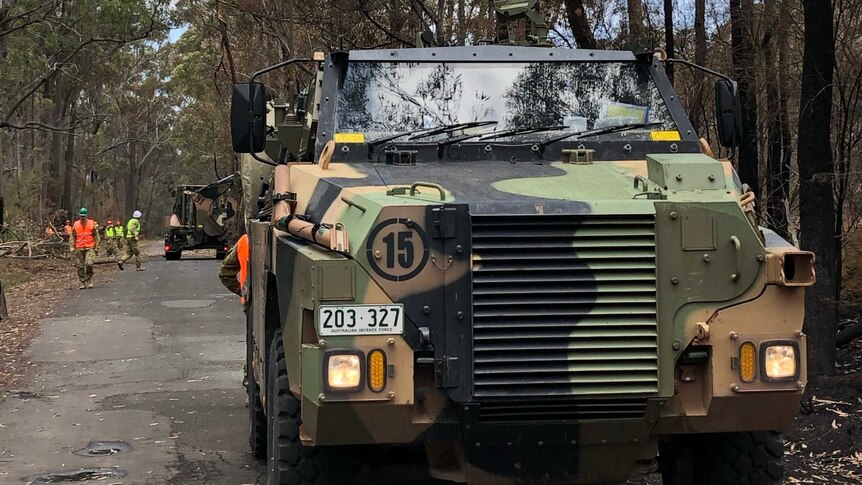 An army vehicle in the foreground with workers clearing trees off the road in the background.
