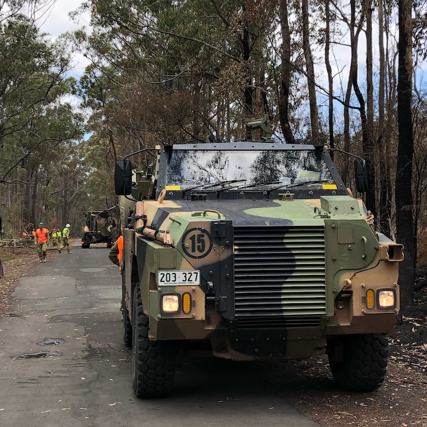 An army vehicle in the foreground with workers clearing trees off the road in the background.