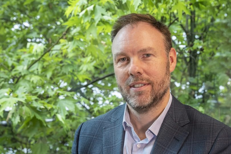 Michael Brennan, a man wearing a dark checked suit and mauve shirt, poses for a photo in front of large green trees