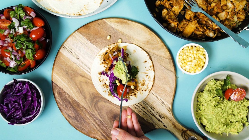 Hand adding guacamole to a barbecue pulled jackfruit taco surrounded by salsa, cabbage, pulled jackfruit and corn tortillas.