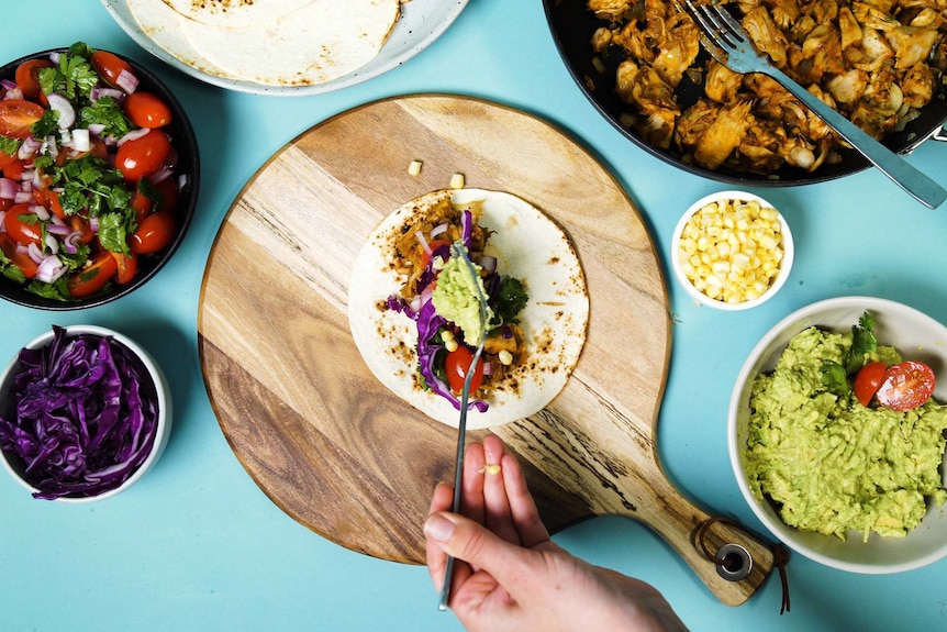 Hand adding guacamole to a barbecue pulled jackfruit taco surrounded by salsa, cabbage, pulled jackfruit and corn tortillas.