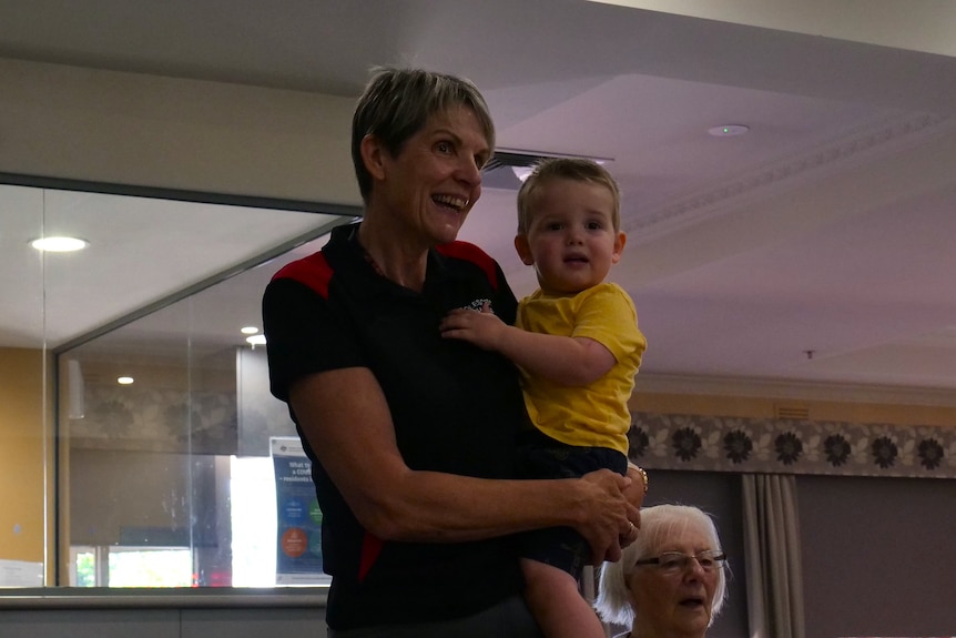 a short-haired woman in a black tshirt is smiling as she holds a toddler in a yellow shirt