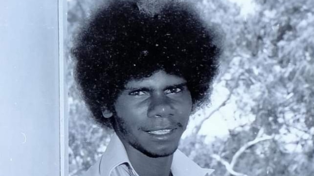 A black and white photo of an Aboriginal man with an afro sitting against a pole. 