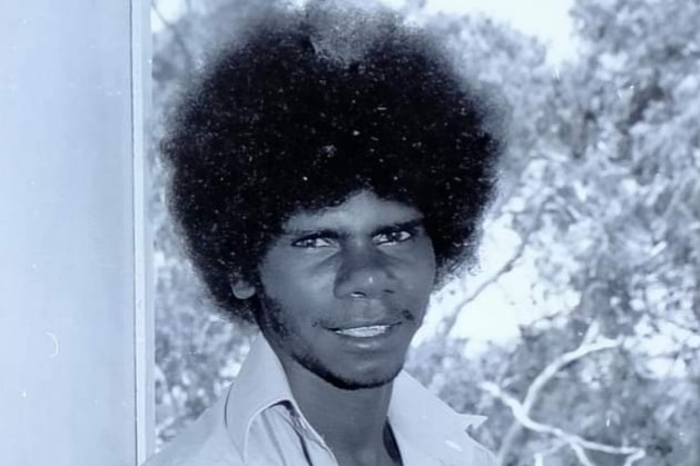 A black and white photo of an Aboriginal man with an afro sitting against a pole. 