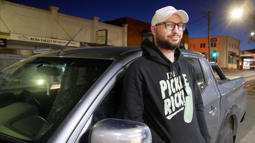 a man wearing a jumper, glasses and cap leans against his car