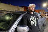 a man wearing a jumper, glasses and cap leans against his car