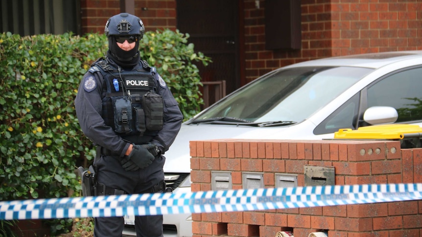 A police officer, in full body protective gear, stands behind police tape outside a red brick house.