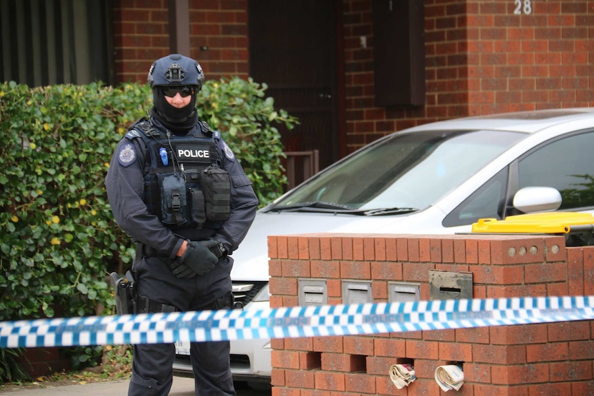 A police officer, in full body protective gear, stands behind police tape outside a red brick house.