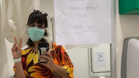 A young woman poses in hospital wearing a face mask and showing the peace sign.