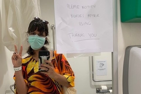 A young woman poses in hospital wearing a face mask and showing the peace sign.