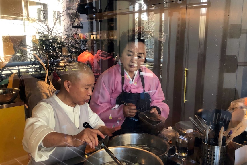 Two women tend to a pot on the stove in a kitchen, as seen through a glass window.