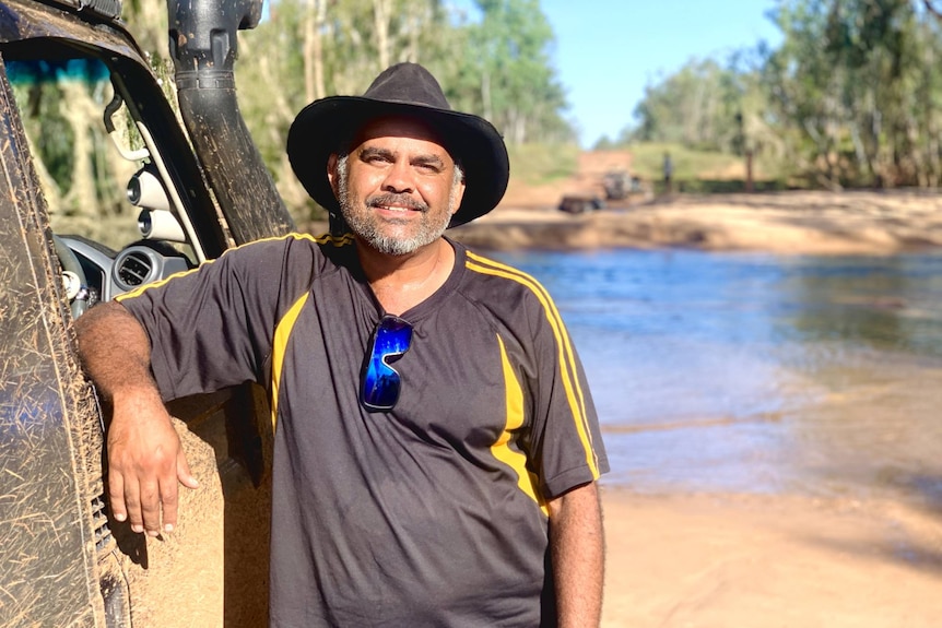 A man stands beside a running river with his elbow on his four-wheel drive.