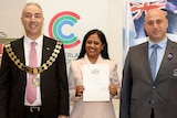 Two formally dressed men, one wearing ceremonial mayoral chains, stand beside a woman who proudly smiles & holds a certificate 