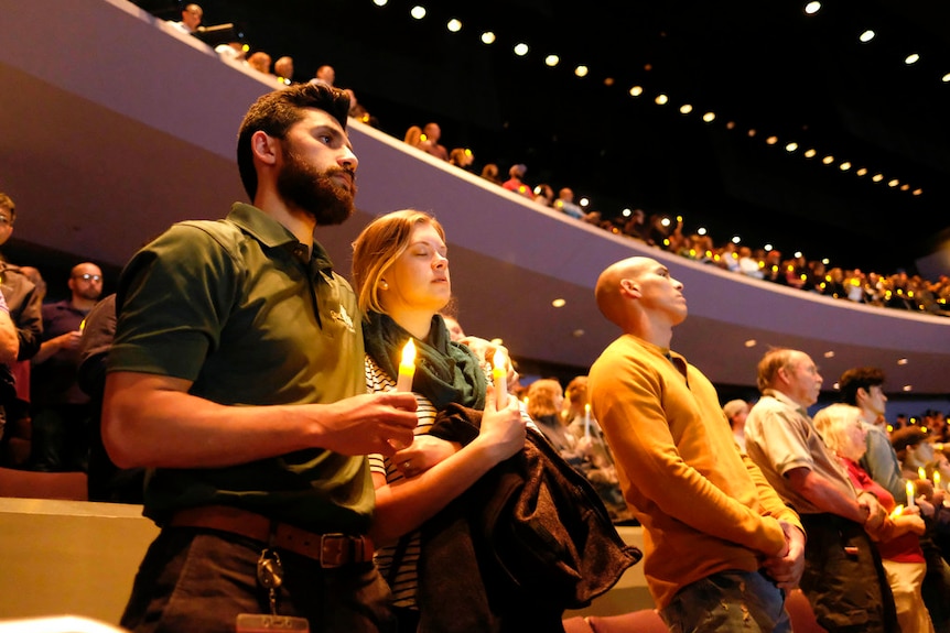 Hundreds of people hold candles and close their eyes as in an auditorium