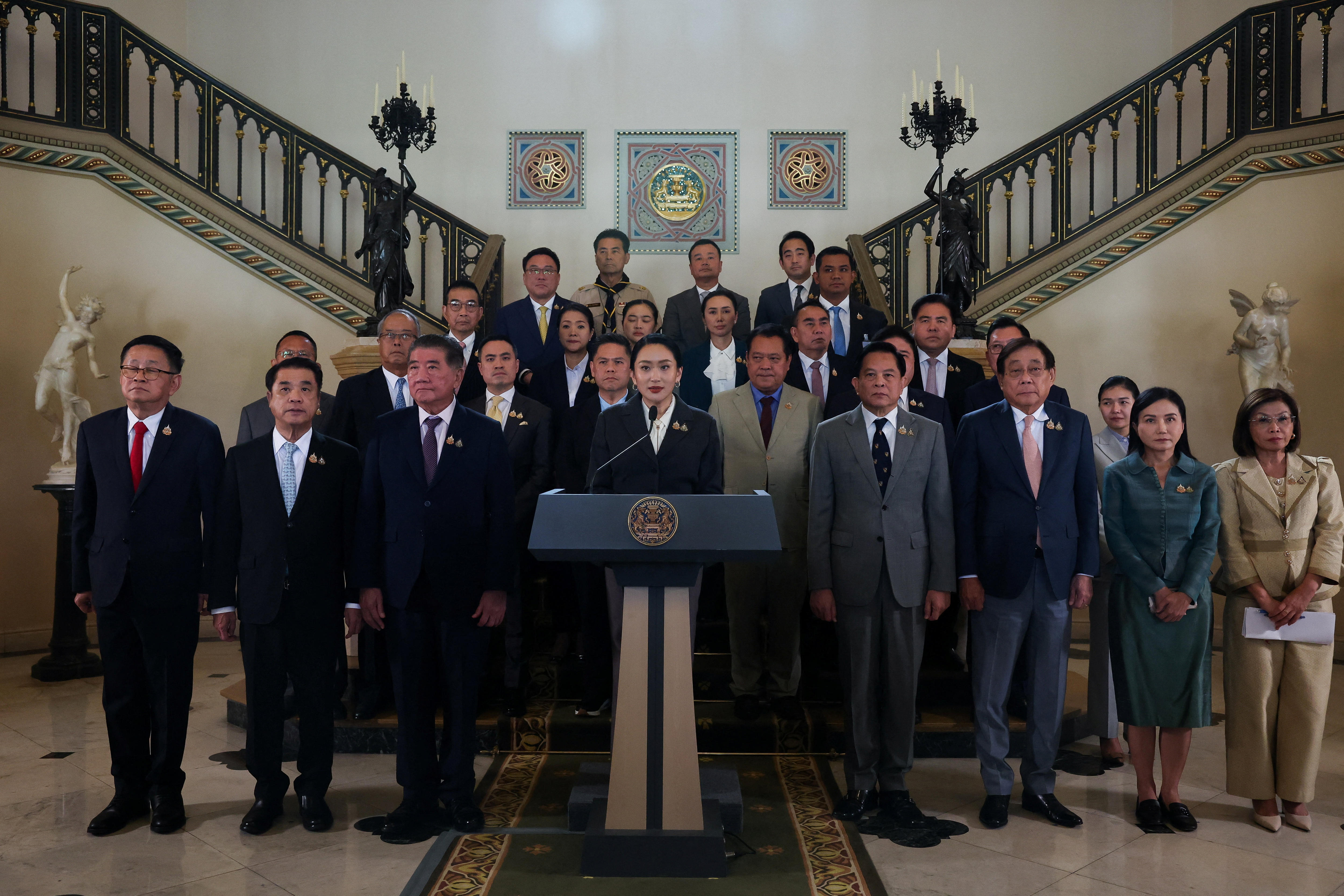 A female with dark hair speaks into a microphone, surrounded by dozens of people in suits 