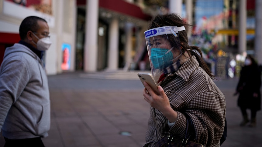 A woman wearing a blue protective mask and face shield looks at her phone while walking past other pedestrians